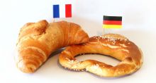 Brezel mit Deutschland-Fähnchen und Croissant mit Frankreich-Fähnchen vor hellem Hintergrund