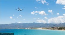 Flugzeug fliegt über Küstenlandschaft mit blauem Meer, blauem Himmel und bewaldeten Bergen