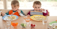 Zwei kleine Kinder mit Latz essen von bunten Tellern zu Mittag, am Tisch sitzend mit Rohkost in Gläsern und Wassergläsern dabei