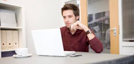 junger Mann in rotem Hemd schaut in Büro auf weißen Laptop und hat eine SCheckkarte in der Hand