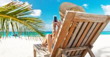 Person mit Smartphone im Liegestuhl am Sandstrand mit blauem Himmel und einem Palmenzweig