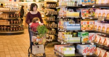 Mutter mit Kind im Einkaufswagen läuft im Supermarkt an Regalen mit Cerealien vorbei