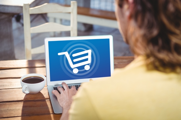 Langhaariger Verbraucher sitzt auf Terrasse mit Tasse Kaffee und hat auf seinem Laptop einen stilisierten Einkaufswagen min weiß mit blauem Hintergrund