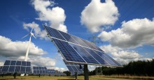 Solarpaneele auf einer Wiese montiert unter blauem Himmel mit weißen Wolken, im Hintergrund  ein Windrad