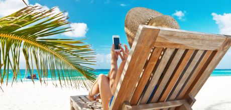 Urlauberin mit Strohhut und Smartphone in der Hand (von hinten) fläzt an einem Palmenstrand mit weißem Sand und blauem Meer