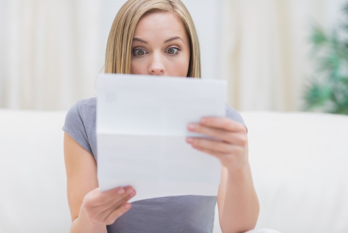 Jüngere Frau sitzt auf dem Sofa und hält mit erstauntem Gesicht ein Blatt Papier vor sich