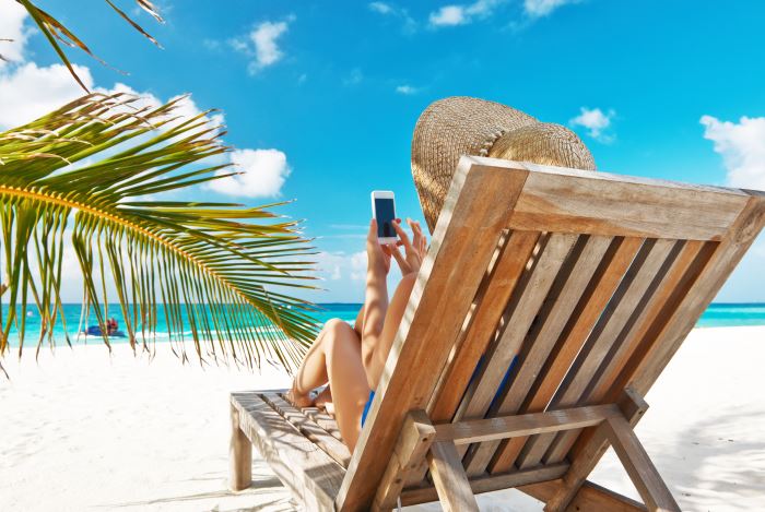 Rückansicht einer Urlauberin am Strand neben Palmwedel auf Holzliegestuhl mit Strohhut auf dem Kopf und Smartphone in der Hand, dazu blauer Himmel