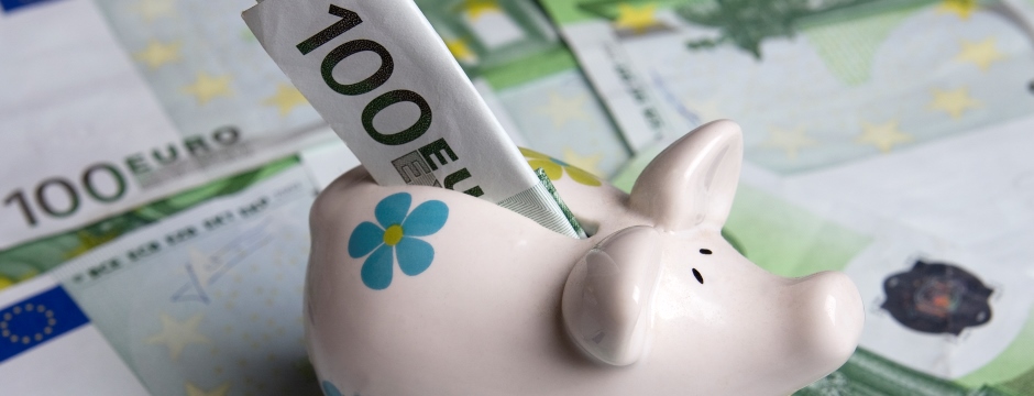 rosa Sparschwein mit blauem Blümchen und 100-Euro-Schein im Schlitz steht auf weiteren 100-Euro-Scheinen