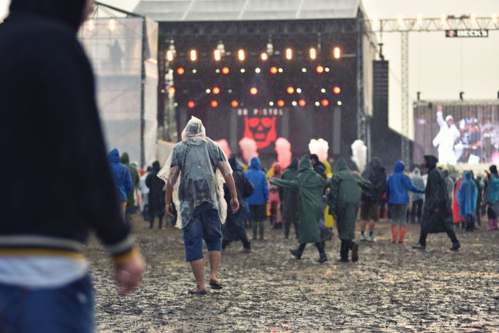 Matschiges Festgelände, der Blick zur beleuchteten Bühne zeigt Festivalbesuche in bunten Regenkleidern auf Matschr