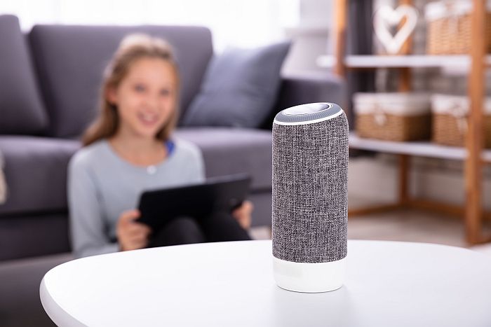 Ein Mädchen mit langen Haaren steuert vor einem Sofa im Hintergrund einen smarten Lautsprecher im Vordergrund. Ein Mädchen mit langen Haaren steuert vor einem Sofa im Hintergrund einen smarten Lautsprecher im Vordergrund.