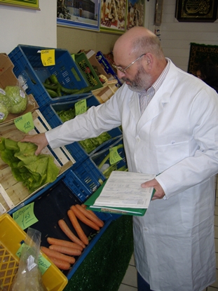 Bärtiger Probennehmer mit Brille in weißem Laborkittel prüft verschiedene Gemüsesorte, Karotten, Salate