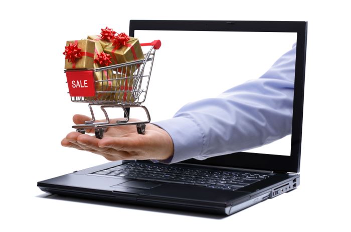 Aus einem Bildschirm eines Laptops ragt eine offene Hand, auf der ein kleiner Einkaufswagen, der die Aufschrift Aus einem Bildschirm eines Laptops ragt eine offene Hand, auf der ein kleiner Einkaufswagen, der die Aufschrift