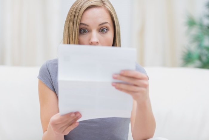 Jüngere Frau sitzt auf dem Sofa und hält mit erstauntem Gesicht ein Blatt Papier vor sich Jüngere Frau sitzt auf dem Sofa und hält mit erstauntem Gesicht ein Blatt Papier vor sich